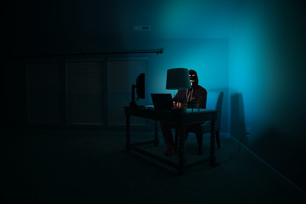 A person in a mask sitting behind a computer in a dimly lit room
